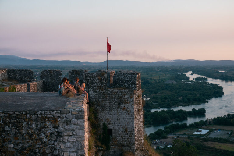 3 people sit on the stone walls of Rozafa Fortress at sunset, looking out over the winding river 