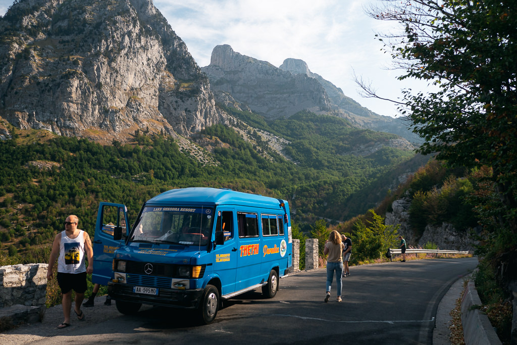 A blue vehicle and people on the road with the mountains in the background in Albania