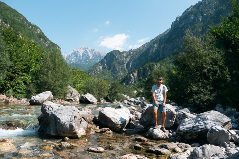 Hiker sits on a boulder in a shallow river within green valleys and peaks of the Albanian Alps