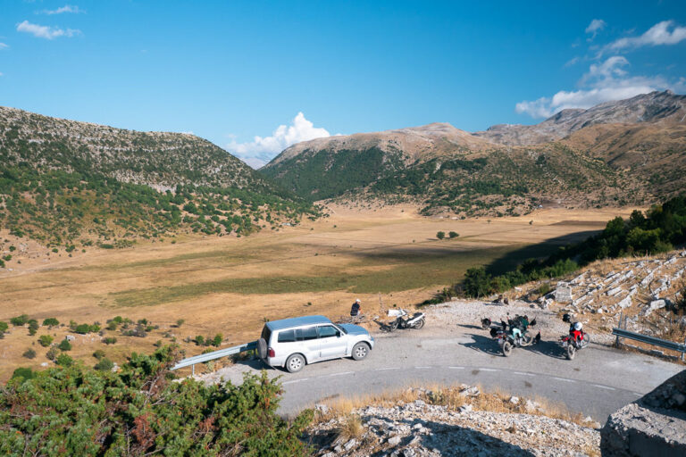 A dry valley in Gjirokastër, with a winding road, a silver SUV, three bikes, and two people by the roadside.