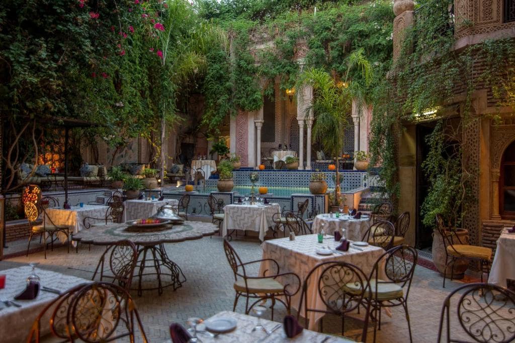 A dining area, with a pool, tables, and chairs, surrounded by plants in a riad in Marrakech