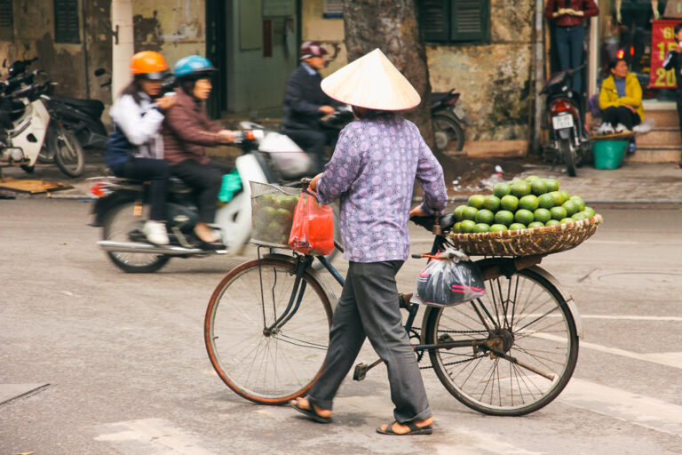 A person wearing a conical hat pushes a bicycle loaded with a basket of fruit in Hanoi