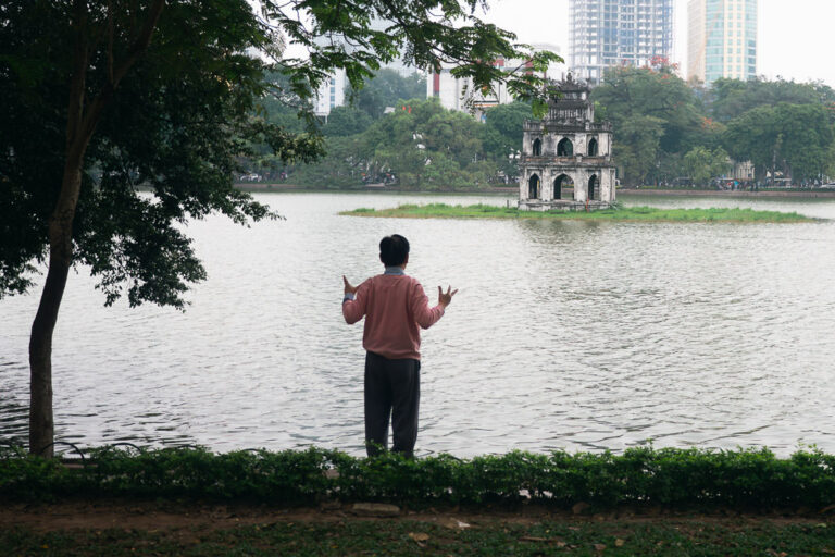 A person stands by the edge of a lake with their arms raised, facing the Turtle Tower (Tháp Rùa) landmark in the center of Hoàn Kiếm Lake in Hanoi