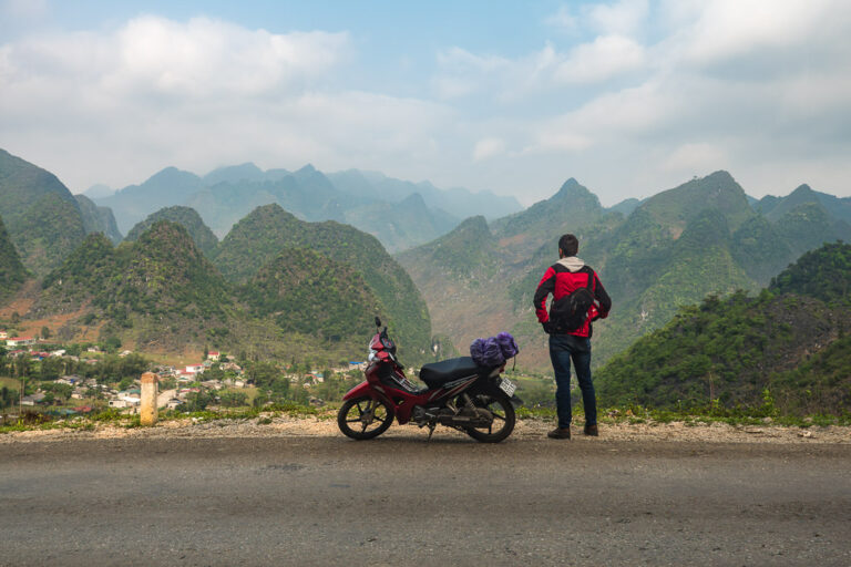 Marek standing next to a red motorbike on a paved road, looking out mountain landscape of Ma Pi Leng Pass