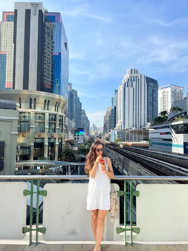 A woman sipping on a drink while waiting on a train in Bangkok, Thailand with buildings in the background.