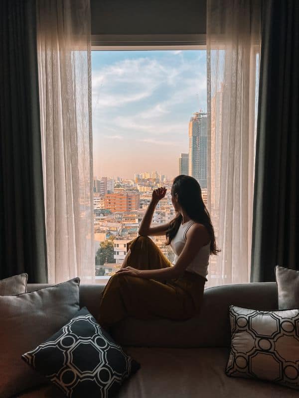 A woman sitting on a sofa inside a hotel room and looking at the view of the Bangkok city view through the window.