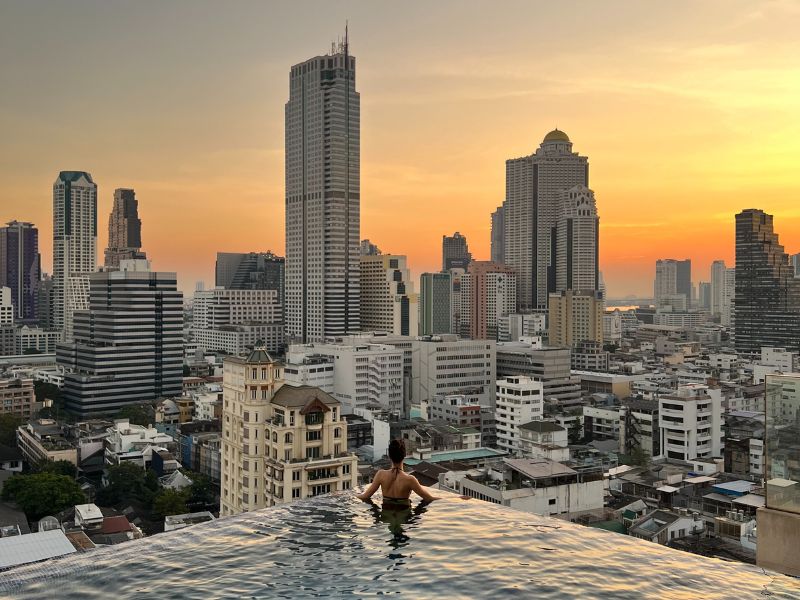 A woman swimming in an outdoor rooftop pool of a hotel while looking at the city buildings at dusk.