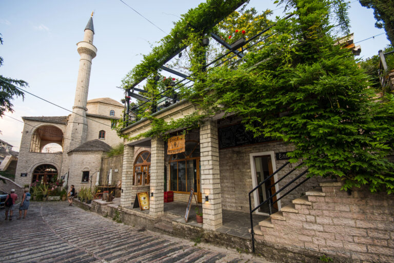 A cobblestone street lined with a vine-covered restaurant, set against Gjirokastër’s historic Bazaar Mosque