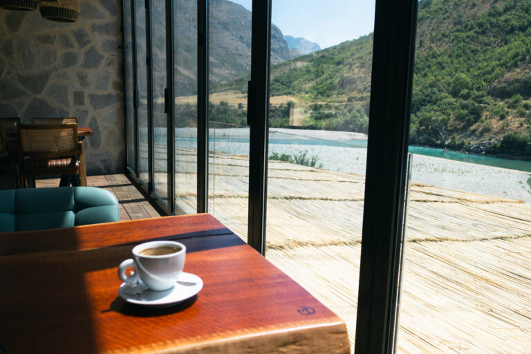 coffee cup on a wooden table, set against a floor-to-ceiling glass wall with a view of the wild Vjosa River