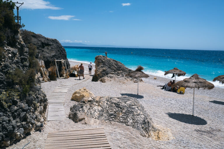 Sunny day at Livadi Beach, Himarë, with a walkway to the pebble shore and people by the sea