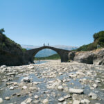 People walking over the Kadiut Bridge crossing a rocky riverbed in Langarica Canyon