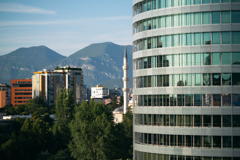 A curved glass office building dominates the right foreground, partially obscuring the view of the Tirana skyline