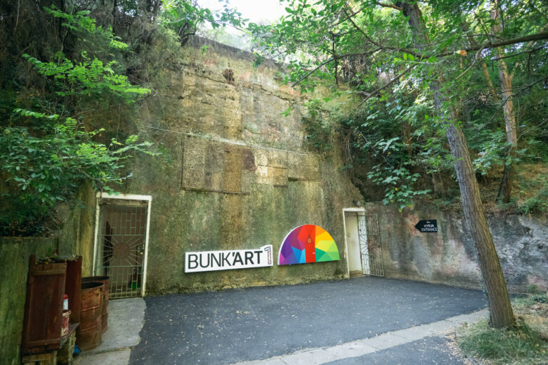 The exterior entrance to Bunk'Art 1 Museum in Tirana with a mossy wall and lush trees and foliage