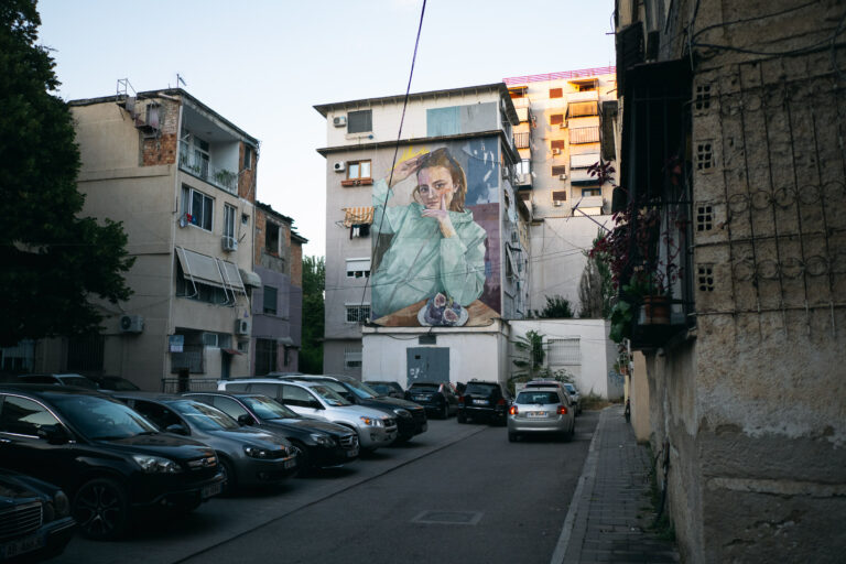 A large-scale mural of a young woman is painted on the side of a five-story apartment building in Tirana