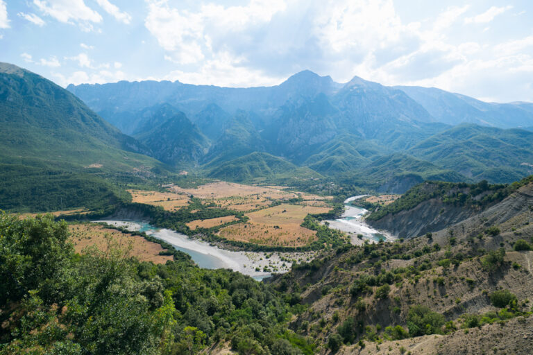 Vjosa River winding through fields in Permet, framed by hills and distant hazy mountains