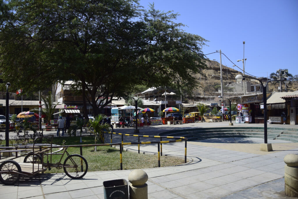 Sunlit town square in Máncora, Peru, with a big tree, a parked bicycle, colorful vendor stalls, and a hillside in the background