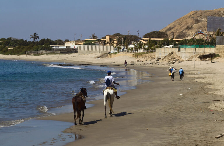 Máncora beach with two horseback riders on wet sand, turquoise ocean, and a village at the base of a sandy hill