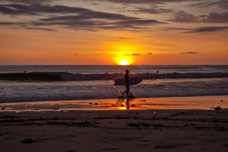 a surfer holding a board and walking by a beach at sunset in Santa Teresa, Costa Rica