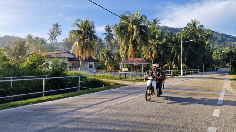 A delivery rider on a motorcycle driving along a paved road in the countryside of Balik Pulau, Penang