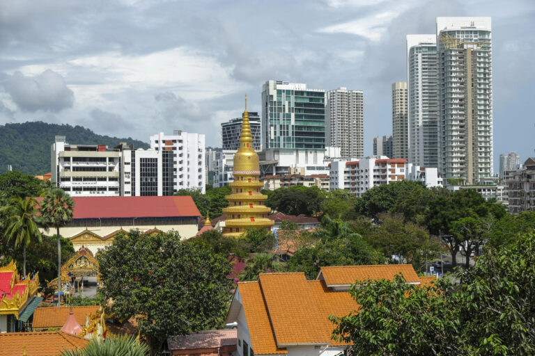 A tiered golden pagoda at Wat Chaiyamangalaram surrounded by trees and modern city buildings