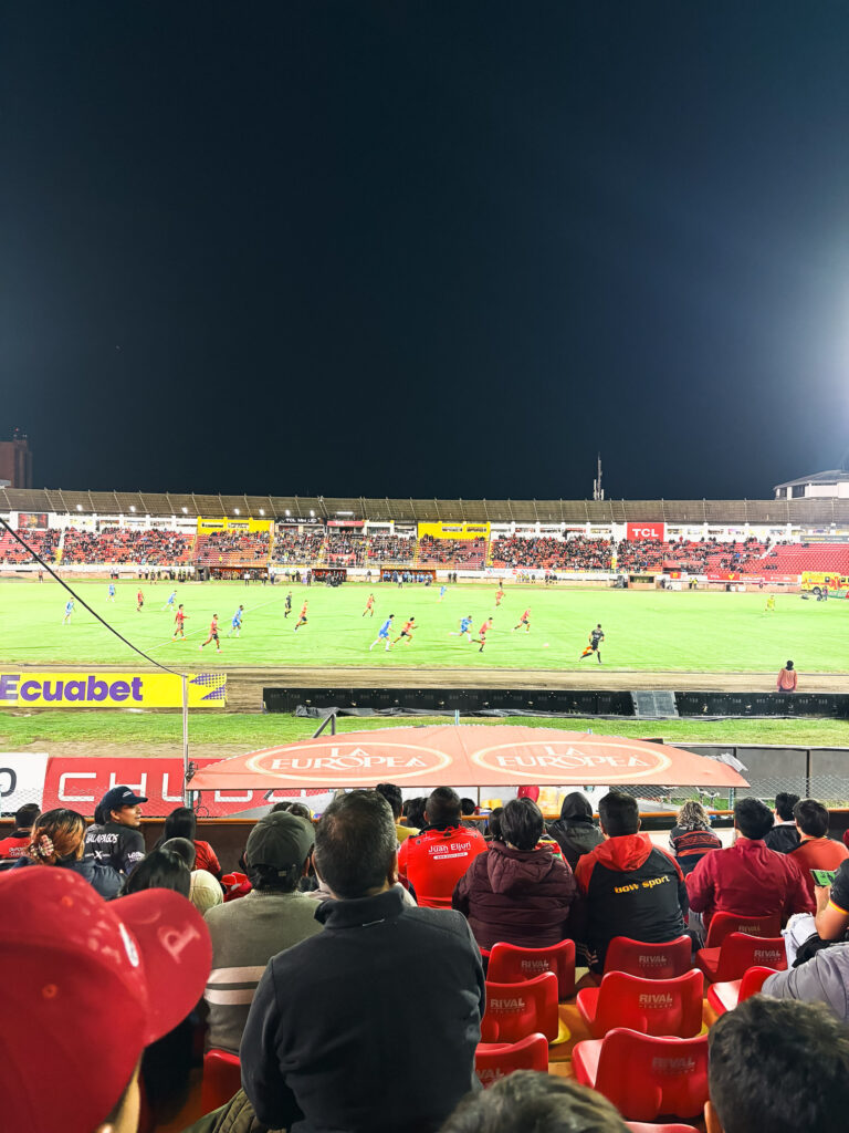 Fans sitting on red seats attending a night football match at Estadio Alejandro Serrano Aguilar in Cuenca